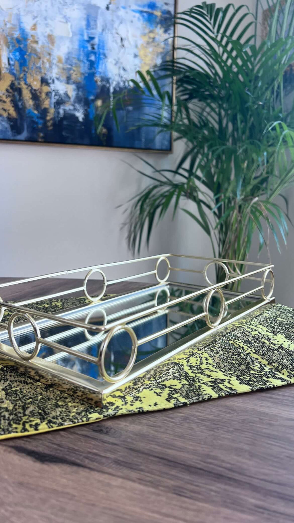 Gold Rectangular Mirror Tray Loop Design displayed on a table next to a green plant and colorful artwork.